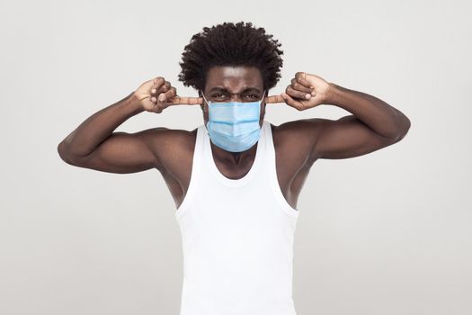 I dont want to hear more. Portrait of tired young man wearing white shirt with surgical medical mask standing and putting his finger on ears and angry. indoor studio shot isolated on gray background.