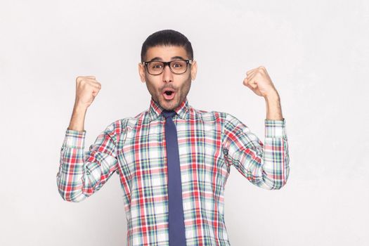 Happy winner handsome bearded businessman in checkered shirt, blue tie and black eyeglasses standing shouting and looking at camera with raised hands. indoor studio shot, isolated on grey background