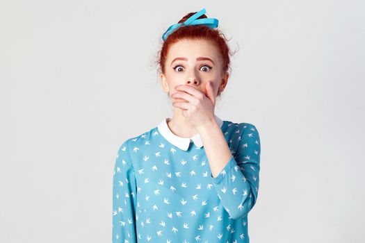 Female in despair and shock. Portrait of young desperate redhead girl in blue dress looking panic, covered her mouth by hand. Isolated studio shot on gray background.