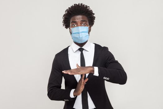 I need more time. Portrait of worry young worker man wearing black suit with surgical medical mask standing with timeout gesture and looking at camera. indoor studio shot isolated on gray background.