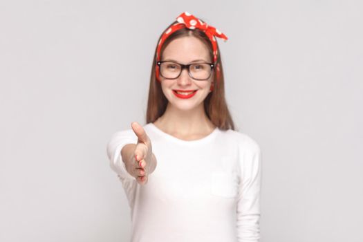 hi, nice to meet you. handshake portrait of beautiful emotional young woman in white t-shirt with freckles, black glasses, red lips and head band. indoor studio shot, isolated on light gray background