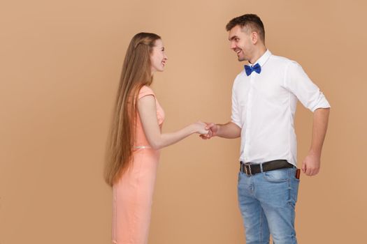 Nice to meet you. Profile side view handshake of handsome man in white shirt and long hair blonde woman in pink dress looking with toothy smile. indoor studio shot, isolated on light brown background
