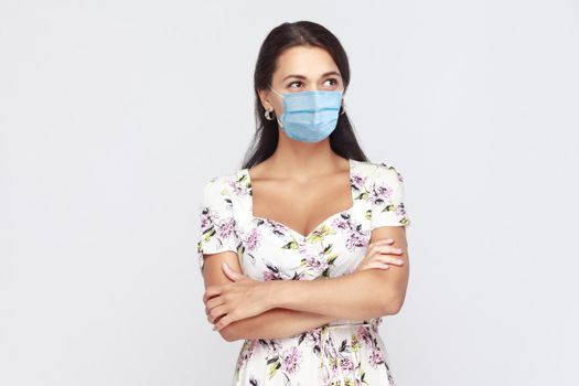 Portrait of thoughtful young brunette woman with surgical medical mask in white dress standing, crossed arms, and looking away and thinking what to do. indoor studio shot isolated on gray background.