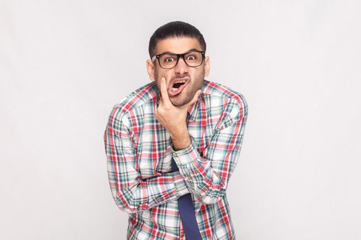 Portrait of сrazy shocked bearded businessman in colorful checkered shirt, blue tie and eyeglasses standing touching face and looking at camera. indoor studio shot, isolated on light grey background.