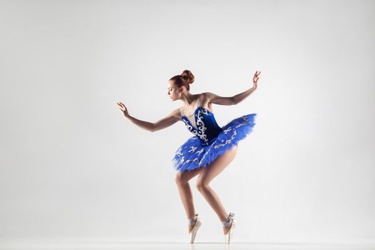 attractive ballerina with bun collected hair wearing blue dress and pointe shoes performing in white studio. indoor, studio shot.