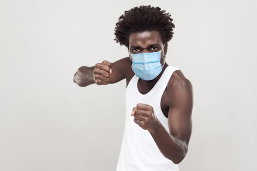 Portrait of angry young man wearing white shirt with surgical medical mask standing with boxing fists and ready to attack or defence. indoor studio shot isolated on gray background.