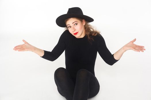 Portrait of confused woman in hat, sitting on floor. Studio shot, isolated on white background