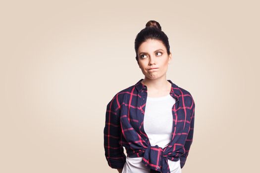 beautiful girl looking away, having doubtful and indecisive face expression, pursuing her lips as if forbidden to say anything. Confused young female posing. studio shot on beige background.