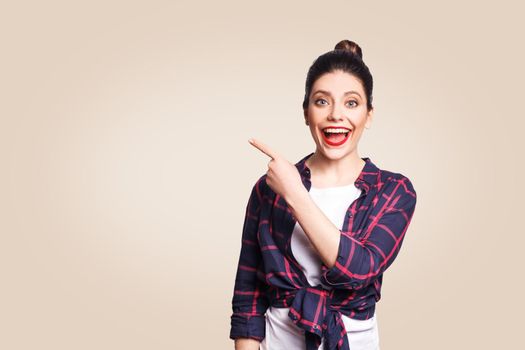 Young happy girl with casual style and bun hair pointing her finger sideways, demonstrating something on beige blank wall with copy space for your information or promotional content.