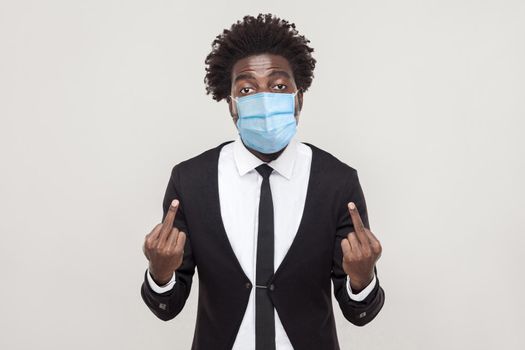 Go away from here Portrait of angry young man wearing black suit with medical mask standing and looking with aggressive face and showing middle finger. indoor studio shot isolated on gray background.