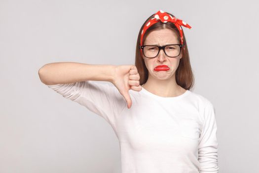 its bad, sad thumbs down of unsatisfied emotional young woman in white t-shirt with freckles, black glasses, red lips and head band looking at camera. studio shot, isolated on light gray background.