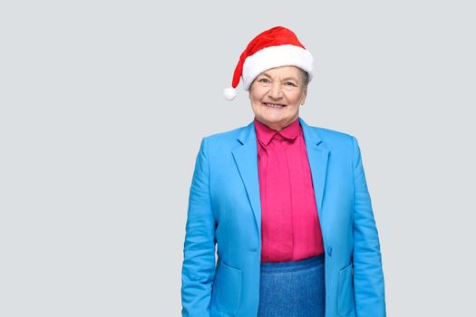 Satisfied toothy smiling colorful casual style aged woman with blue suit and christmas santa red cap standing and looking at camera with joyful face. indoor, studio shot, isolated on gray background