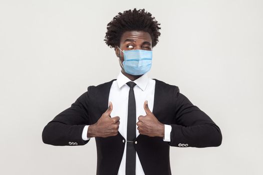 Like. Portrait of satisfied funny young hamdsome worker man wearing black suit with surgical medical mask standing, thumbs up and looking away. indoor studio shot isolated on gray background.