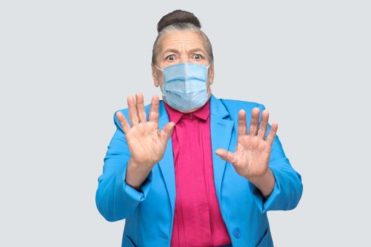 aged woman with surgical medical mask scared, have fear face. Grandmother with light blue suit and pink shirt standing with collected bun gray hair. indoor studio shot, isolated on gray background