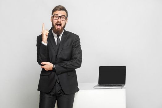 New idea! Portrait of handsome suprised bearded young businessman in black suit are standing near his working place and showing finger up with open mouth. Isolated,studio shot, indoor, gray background