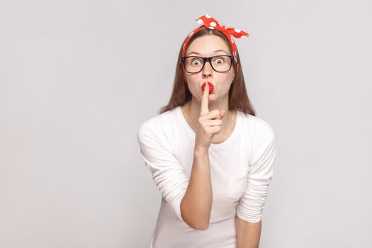 shh! this is secret. portrait of beautiful emotional young woman in white t-shirt with freckles, black glasses, red lips and head band. indoor studio shot, isolated on light gray background.