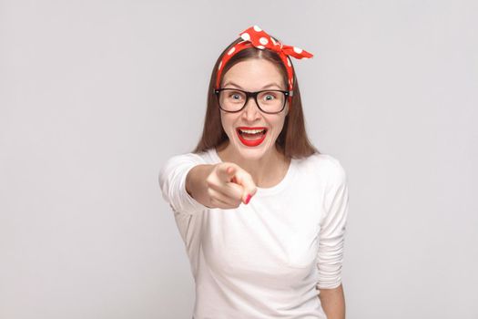 its it you? pointing finger at camera. portrait of beautiful emotional young woman in white t-shirt with freckles, black glasses, red lips and head band. indoor, isolated on light gray background.