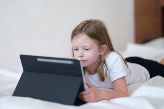 Little girl carefully looks at tablet while lying on bed. Mobile learning apps for kids concept