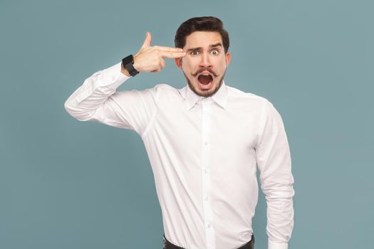 crazy young businessman with shocked face showing gun sign. Suicide concept. portrait of sad bearded businessman in white shirt, with smart watch. Indoor studio shot, isolated on light blue background