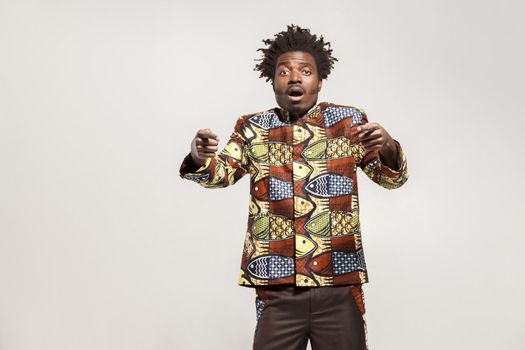 Amazed afro man pointing fingers at camera and shock. Indoor, isolated on gray background