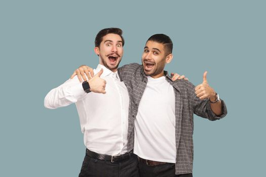 Portrait of two handsome bearded happy friends or partners standing hugging, and looking at camera with satisfied amazed face and thumbs up. indoor studio shot, isolated on light blue background.