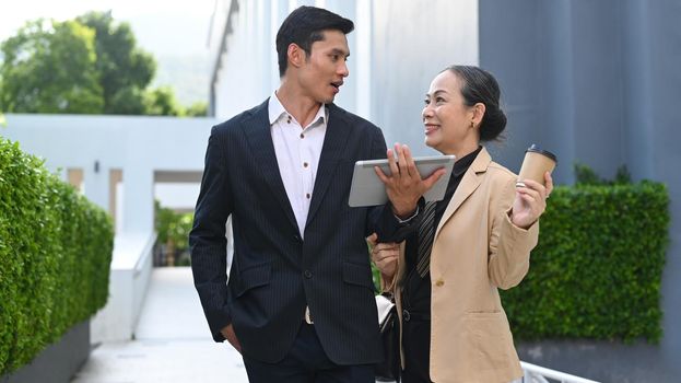 Smart young asian businessman discussing business plan with senior businesswoman while walking outside modern office building.