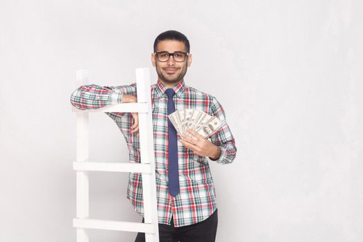 Portrait of successful rich handsome bearded young man in colorful checkered shirt with blue tie standing, holding fan of cash and lean on white stairs. Indoor studio shot, isolated on grey background