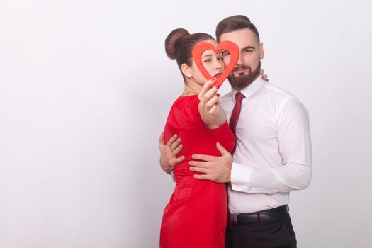 Beautiful couple embrace with passion, holding red heart. Indoor, studio shot, isolated on gray background
