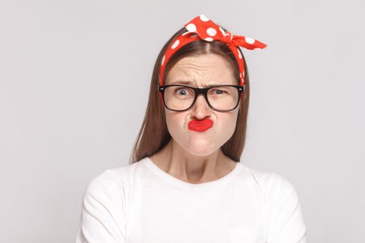 crazy wondered funny portrait of beautiful emotional young woman in white t-shirt with freckles, black glasses, red lips and head band. indoor studio shot, isolated on light gray background.