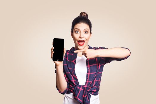 Young beautiful shocked woman in casual style holding phone looking at camera and showing phone display. studio shot on beige background.