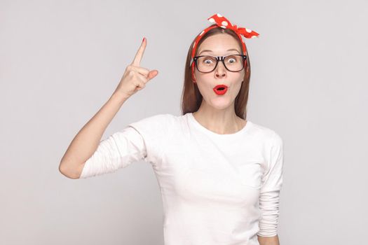 wow. I have an idea. portrait of beautiful emotional young woman in white t-shirt with freckles, black glasses, red lips and head band. indoor studio shot, isolated on light gray background.