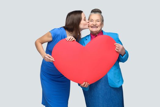 Granddaughter kissing grandmother standing, holding big heart shape, looking at camera with toothy smile. Big love in family. Relations in the family. indoor studio shot, isolated on gray background