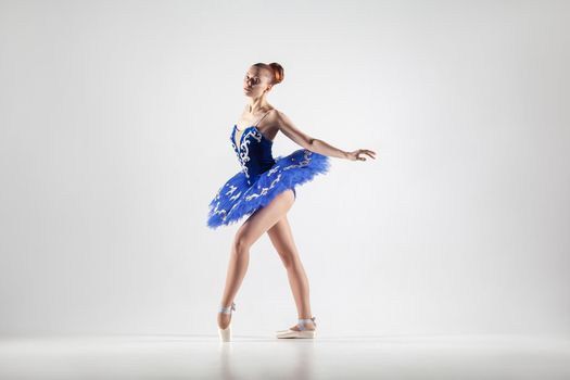 attractive ballerina with bun collected hair wearing blue dress and pointe shoes performing in white studio. indoor, studio shot.