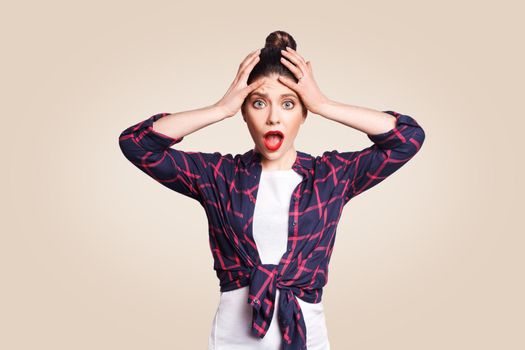 portrait of hysterica girl with black bun hair looking in despair and panic, not knowing what to do, hands on her head, mouth wide open, and big eyes, looking at camera. on beige background.