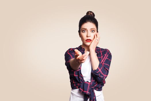 Scared girl with bun hair knot having scared and frightened look, keeping mouth open, touching face and pointing hand at camera.