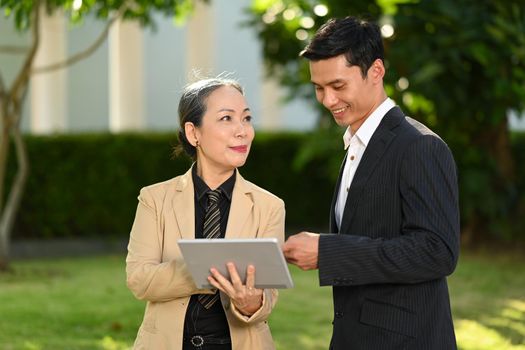 Mature female investor holding digital tablet and explaining business plan to Asian male adviser while standing at modern office district.