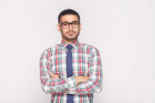 portrait of happy handsome bearded businessman in colorful checkered shirt, blue tie and black eyeglasses standing in crossed hands and smiling. indoor studio shot, isolated on light grey background.