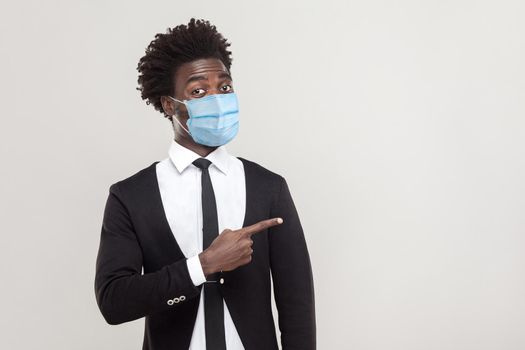 Portrait of funny young handsome worker man wearing black suit with surgical medical mask standing showing and pointing at empty background copyspace. indoor studio shot isolated on gray background.