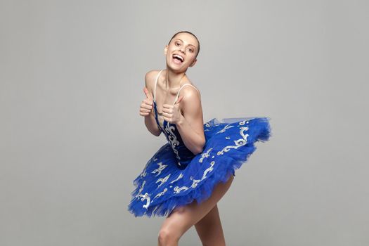Thumbs up. Portrait of satisfied beautiful ballerina woman in blue costume with makeup and bun collected hairstyle standing against gray background. emotion and expression concept. indoor studio shot.