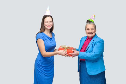 Grandmother in blue suit and hat holding red gift box and giving to her lovely granddaughter on her birthday. family happiness life event celebration. indoor studio shot, isolated on gray background