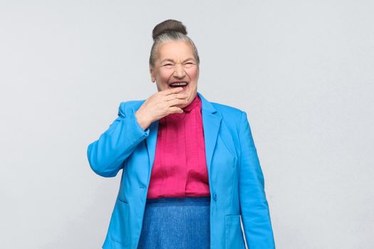 Funny aged woman laugh . Emotion and feelings. Portrait of handsome expressive grandmother with light blue suit and pink shirt standing with collected bun gray hair. indoor isolated on gray background