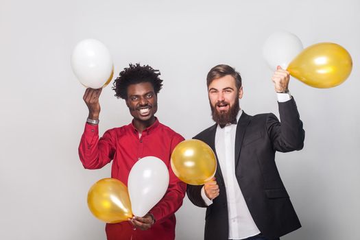 Happiness friends celebrating birthday, holding gold and white air balloon, hands up and smiling. Studio shot, gray wall