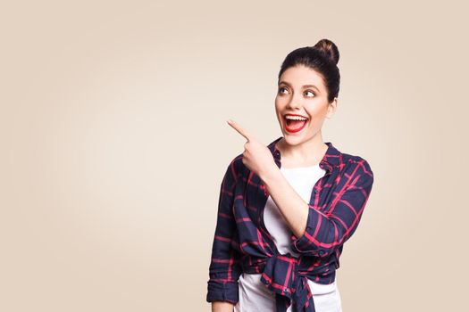 Young happy girl with casual style and bun hair pointing her finger sideways, demonstrating something on beige blank wall with copy space for your information or promotional content.