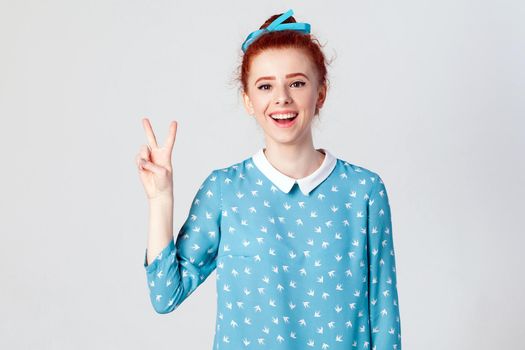 Happiness beautiful redhead girl shows peace or victory sign. Isolated studio shot on gray background.