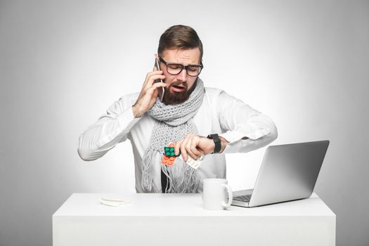 Confused cold sick young msn in white shirt and scarf are sitting in office on desk and talking with partner on phone also checking time on his own hand watch, planning meeting. indoor gray background