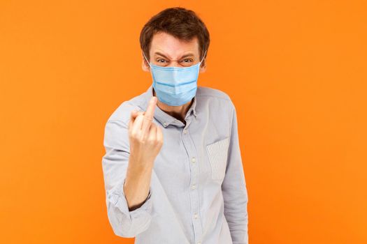 Portrait of angry young worker man with surgical medical mask standing looking at camera with middle finger and aggressive face. indoor studio shot isolated on orange background.