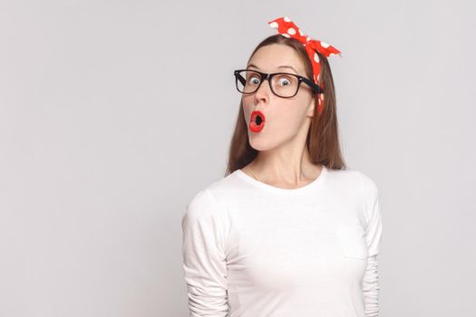 wow its unbelievable, portrait of beautiful emotional young woman in white t-shirt with freckles, black glasses, red lips and head band. indoor studio shot, isolated on light gray background.