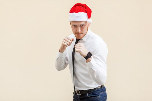 Boxing. Young adult ginger businessman on red santa hat, ready for fight on light orange background. Studio shot