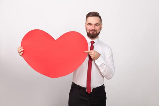 Perfect businessman smiling, pointing finger at big red heart. Indoor, studio shot, isolated on gray background