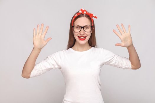 hey, nice to see you. portrait of beautiful emotional young woman in white t-shirt with freckles, black glasses, red lips and head band. indoor studio shot, isolated on light gray background.